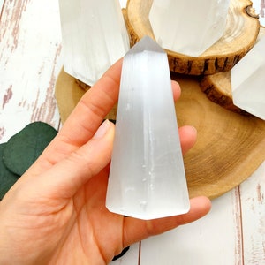 Könnte beinhalten: Ein weißer Selenit-Kristallspitz, auch bekannt als Selenit-Stab, wird in einer Hand gehalten. Der Kristall ist glatt und hat eine durchscheinende Optik. Der Kristall liegt auf einer Holzoberfläche.