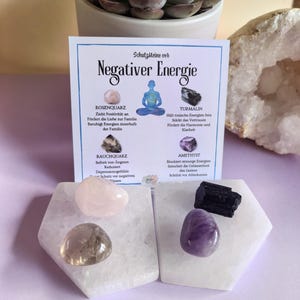 Könnte beinhalten: Ein Set aus vier Kristallen auf weißen Ständern mit einer Karte, die ihre Eigenschaften beschreibt. Die Kristalle sind Rosenquarz, Rauchquarz, Amethyst und schwarzer Turmalin. Die Karte lautet "Schutzsteine vor Negativer Energie", was übersetzt "Schutzsteine gegen negative Energie" bedeutet.