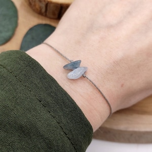 Könnte beinhalten: Ein graues Armband aus Schnur mit zwei hellblauen Stein-Anhängern. Das Armband ist an einem Handgelenk gebunden.