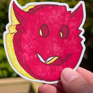 May include: A die-cut sticker featuring a stylized red devil face with yellow horns and a yellow tongue. The devil has a textured, watercolor-like appearance with a dark red outline, yellow eyes, and a wide, mischievous grin showing a yellow tongue.