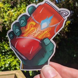 May include: A die-cut sticker featuring a green-skinned hand holding a red soda can. The can displays the word "Sprite" in yellow cursive lettering with a blue flame icon above it. The design includes white highlights to simulate condensation on the can.