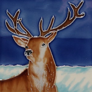 Könnte beinhalten: Eine Keramikfliese mit einem handgemalten Bild eines braunen Hirsches mit großen Geweih auf blauem Hintergrund. Der Hirsch steht auf einer schneebedeckten Wiese.