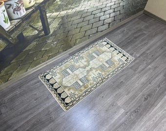 May include: A small, beige rug with a geometric pattern in black and white. The rug has a fringed edge and is laid out on a wooden floor.