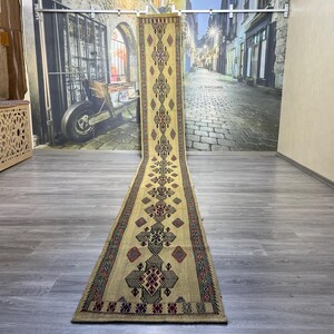 May include: A long, beige runner rug with a geometric pattern in black, red, and blue. The rug has a traditional design with repeating diamond shapes and intricate details.