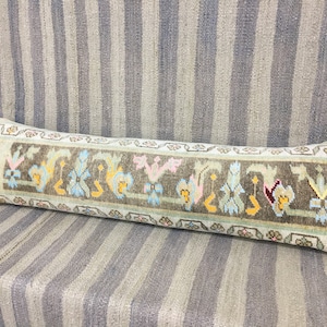 May include: A long, rectangular pillow with a light blue background and a brown, green, yellow, pink, and red patterned border. The pillow is made of woven fabric and has a traditional design.