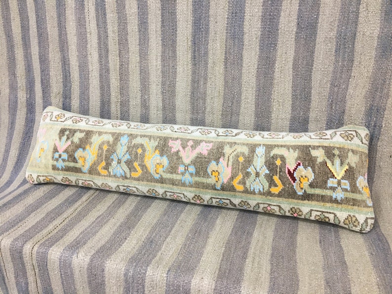 May include: A long, rectangular pillow with a light blue background and a brown, green, yellow, pink, and red patterned border. The pillow is made of woven fabric and has a traditional design.