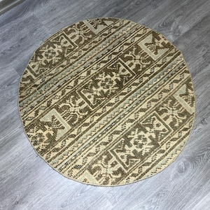 May include: A round rug with a detailed geometric pattern in shades of brown, beige, and light blue. The rug's design features repeating shapes and lines, creating a textured appearance. The rug is placed on a wooden floor.