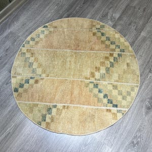 Puede incluir: Alfombra redonda de color beige y melocotón con un diseño geométrico. La alfombra tiene forma circular y está hecha de material tejido. El diseño incluye cuadrados azules y marrones. La alfombra está colocada sobre un suelo de madera.