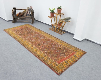 May include: A rectangular, patterned rug with a central field of reddish-brown and a yellow border. The rug features intricate geometric designs and floral motifs. The rug is placed on a gray floor.