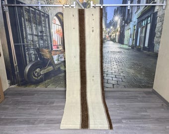 May include: A long, off-white runner rug with two brown stripes running down the center. The rug is hanging from a rod and has a textured, woven look.