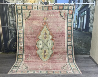 Puede incluir: Una alfombra rectangular grande con una apariencia descolorida y vintage. La alfombra presenta un diseño central en tonos de verde, amarillo y naranja sobre un fondo rosa polvoriento. Un borde verde oscuro con patrones geométricos enmarca la alfombra.
