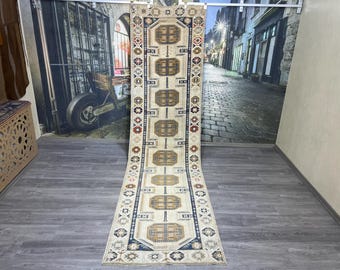 May include: A long, rectangular rug with a cream background and intricate geometric patterns in shades of brown, blue, and red. The rug has a fringed edge and is displayed on a wooden floor.