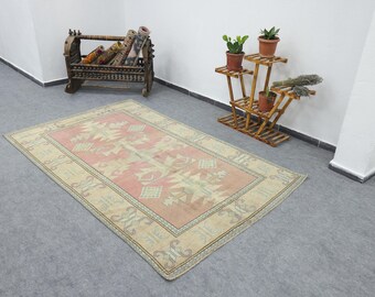 Puede incluir: Una alfombra rectangular con un campo central rosa y un borde beige. La alfombra presenta un patrón geométrico en tonos beige, rosa y verde. La alfombra está colocada sobre un suelo gris.