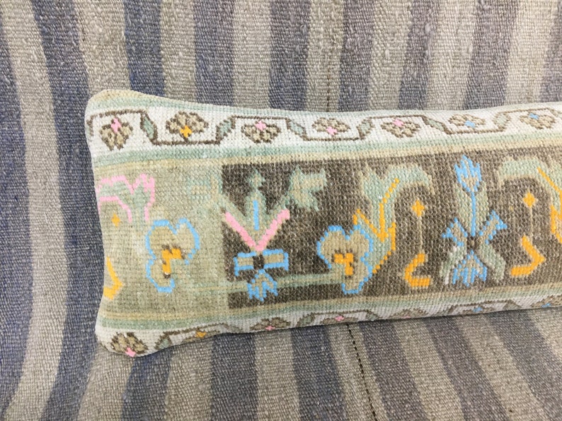 May include: A long, rectangular pillow with a floral pattern in shades of blue, pink, yellow, and brown. The pillow is made of a woven fabric and has a fringed edge.