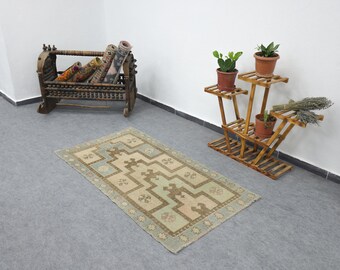 May include: A rectangular rug with a geometric pattern in beige, light blue, and brown. The rug is on a grey floor. A wooden plant stand with potted plants and a wooden cradle with rolled rugs are also in the image.