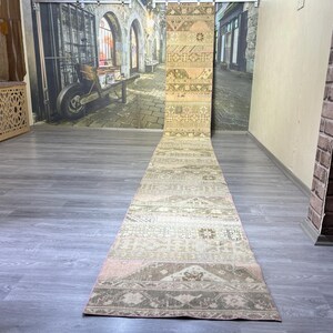 May include: A long, rectangular rug with a faded, vintage appearance. The rug features a patchwork design with various geometric patterns in shades of beige, brown, and pink. The rug is displayed on a gray wooden floor.