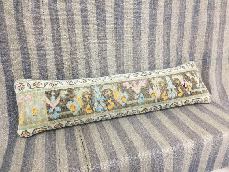 May include: A long, rectangular pillow with a light blue background and a colorful floral pattern. The pillow is made of woven fabric and has a fringe border.