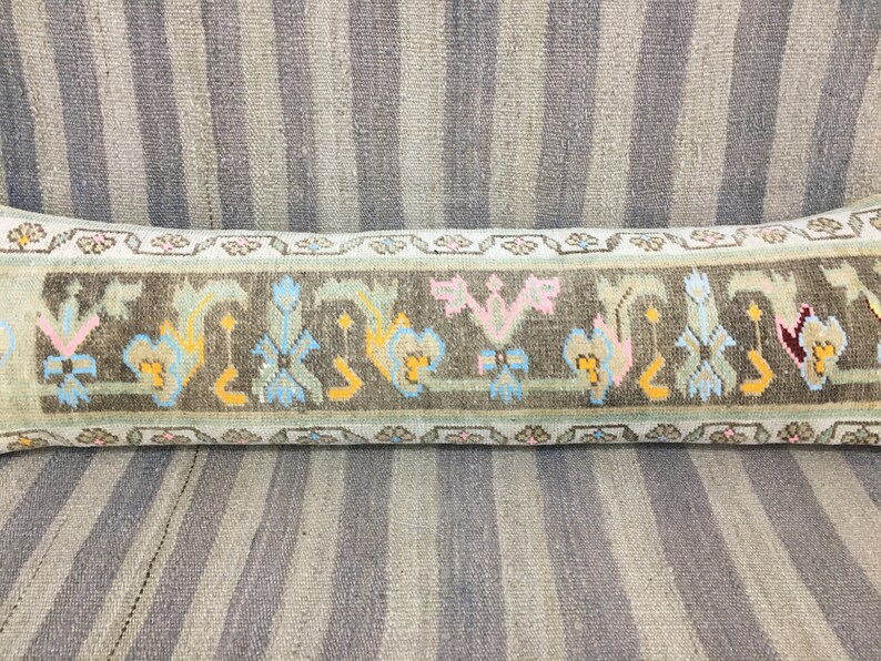 May include: A long, rectangular pillow with a light blue background and a brown, green, pink, and yellow geometric pattern. The pillow is made of woven fabric and has a fringe border.