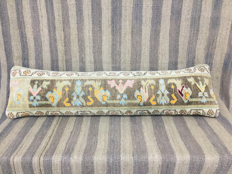 May include: A long, rectangular pillow with a light blue background and a brown, green, and yellow floral pattern. The pillow is made of a soft, woven fabric and has a fringed edge.