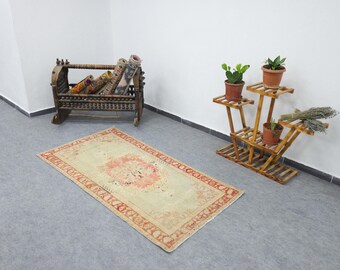 Puede incluir: Una alfombra rectangular beige con un estampado floral rojo y rosa en el centro y un borde rojo. Alfombras enrolladas están en una cuna de madera. Un soporte de madera para plantas sostiene plantas en macetas.
