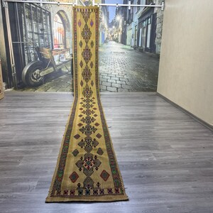 May include: A long, narrow runner rug with a geometric pattern in shades of yellow, brown, red, and blue. The rug has a fringe on both ends.