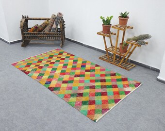 May include: A rectangular, colorful rug with a geometric pattern of squares in shades of red, green, yellow, and orange. The rug is placed on a gray floor, with a wooden plant stand and a decorative wooden rack in the background.