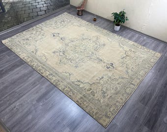 May include: A large, rectangular beige rug with an intricate floral pattern in shades of blue and gray. The rug has a detailed border and a central medallion design. The rug is placed on a dark gray wooden floor.