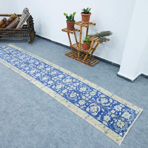 May include: A long, rectangular blue and cream-colored rug with a floral pattern. The rug is placed on a gray floor. A wooden plant stand with potted plants and a decorative wooden cradle are in the background.