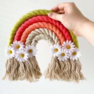 Può includere: Un'arazzo da parete a forma di arcobaleno fatto a mano con una combinazione di colori verde, rosso, arancione e crema. L'arcobaleno è decorato con fiori di margherita bianchi e frange color crema. Una mano tiene l'arazzo su sfondo bianco.