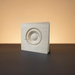 May include: A plain, unfinished wooden rosette with a circular design. The rosette is square and has a smooth surface.