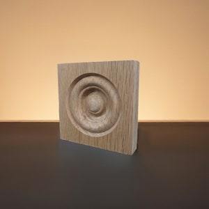 May include: A wooden square block with a carved circular design in the center. The design features concentric circles with a smaller circle in the middle.