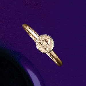 May include: A gold-colored ring with a circular emblem featuring an eye design within a triangle. The ring has a smooth, polished band and a detailed emblem. The background is a deep purple.