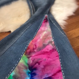May include: Blue denim jeans with a flared design. The jeans feature a vibrant, colorful tie-dye fabric insert in shades of pink, purple, green, and blue. White stitching outlines the insert and the jeans' seams.