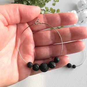 May include: A pair of silver hoop earrings with black lava rock beads. The earrings are shown on a hand, with the beads forming a semi-circle.