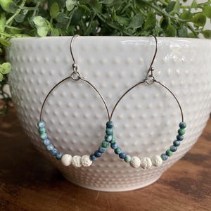 May include: A pair of hoop earrings featuring thin silver-toned metal wires adorned with a sequence of small, round, blue and green marbled beads, accented by three textured white porous beads at the bottom center of each hoop.