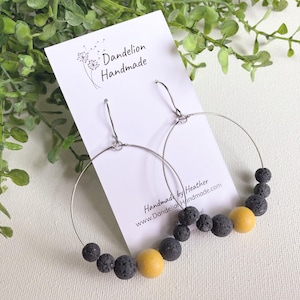 May include: A pair of silver-toned hoop earrings featuring a series of small, porous black lava beads and a single smooth, round yellow bead on each wire. The earrings are displayed on a white card labeled Dandelion Handmade.