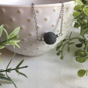 Puede incluir: Un collar de cadena plateada con una cuenta de roca volcánica negra. El collar está apoyado en un cuenco blanco con lunares marrones.