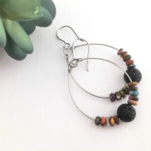 May include: A pair of hoop earrings featuring thin silver-toned metal wires. Each hoop is adorned with a single large, porous black lava bead and several smaller, multi-colored, speckled round beads in shades of blue, orange, and brown.