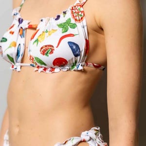 May include: White bikini set with a colorful food-themed print. The top features a ruffled edge and spaghetti straps. The print includes illustrations of red chili peppers, basil leaves, and other food items. The bottoms have tie sides.