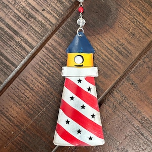 May include: A decorative lighthouse ornament with a red and white striped body and a blue roof. The lighthouse features black star cutouts and is suspended by a silver chain with clear and red beads. The ornament is approximately 13 cm tall.