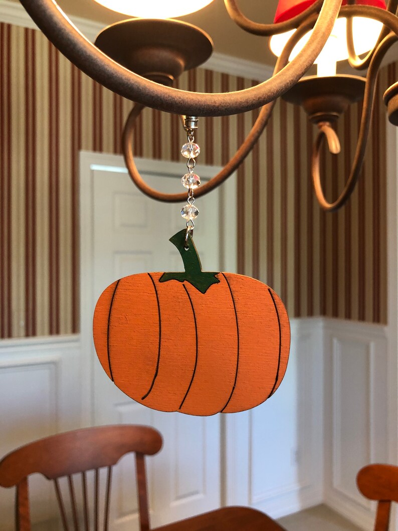 May include: A wooden pumpkin ornament with a green stem and orange paint hangs from a chandelier with a crystal bead chain.