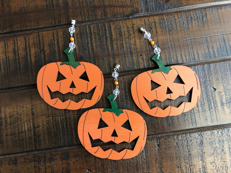 May include: Three orange and green wooden pumpkin ornaments with carved jack-o'-lantern faces. Each pumpkin has a string of clear and orange beads attached to the top.