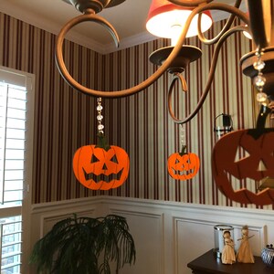 May include: Three orange jack-o'-lantern decorations hang from a chandelier with crystal beads. The decorations are made of paper and have a black outline.