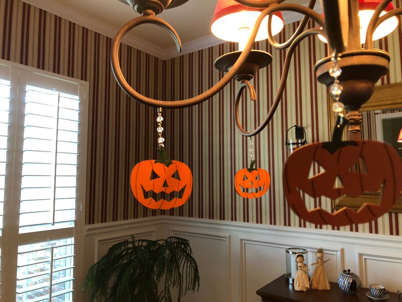 May include: Three orange jack-o'-lantern decorations hang from a chandelier with crystal beads. The decorations are made of paper and have a black outline.