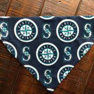 Puede incluir: Un bandana para perro azul y blanco con un patrón repetido del logotipo de los Seattle Mariners. El logotipo presenta una brújula con una pelota de béisbol dentro y las palabras "Seattle Mariners".