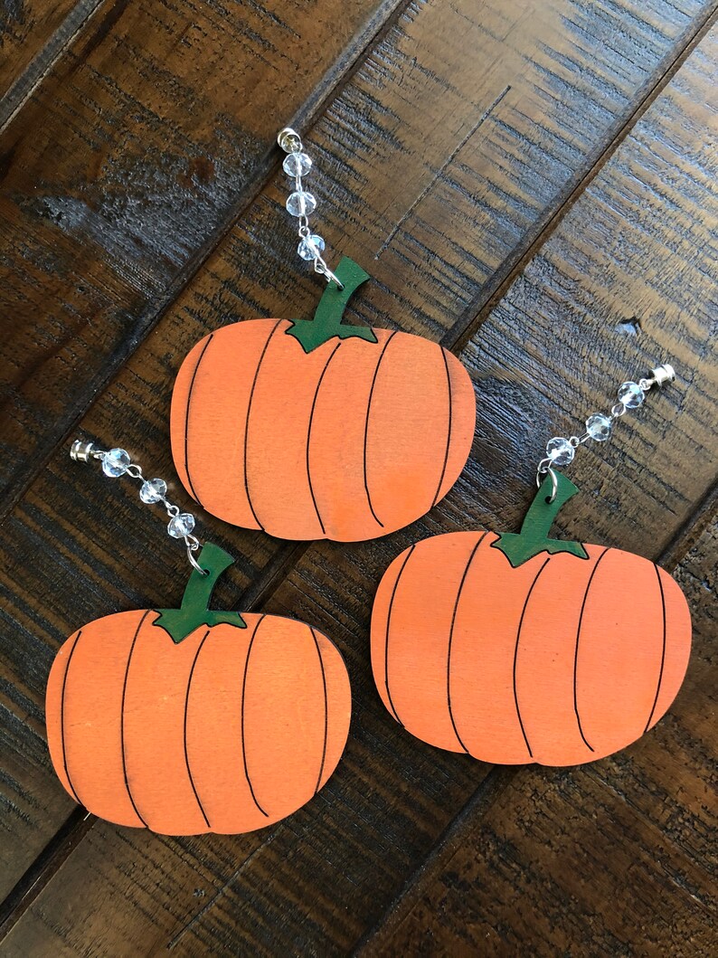 May include: Three orange pumpkin ornaments with green stems and black lines. Each pumpkin has a clear beaded string attached to the top.