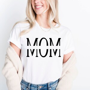May include: White t-shirt with the word "MOM" printed in large, black capital letters. The word is mirrored below itself. The shirt is worn with a beige cardigan and blue jeans.