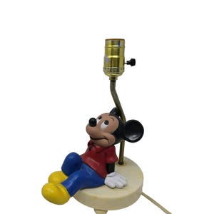 May include: A vintage Mickey Mouse lamp featuring the iconic character in a relaxed pose. The lamp has a red shirt, blue pants, and yellow shoes. The base is off-white with a brass-colored lamp holder.