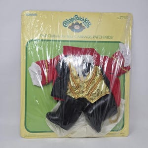 May include: Cabbage Patch Kids doll clothes set, packaged in plastic. The outfit includes a red jacket with gold trim, a gold vest, black pants, and white ruffled sleeves. The packaging is yellow and green with the words "Cabbage Patch Kids" printed on it.