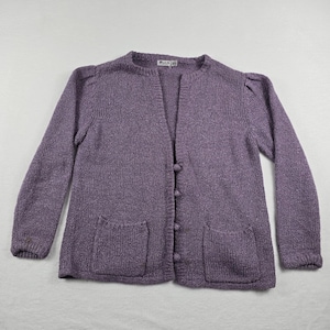 May include: A long-sleeved purple knit cardigan featuring a round neckline, a front button closure with four matching round buttons, and two patch pockets on the lower front. The garment has slightly gathered shoulders and ribbed cuffs.